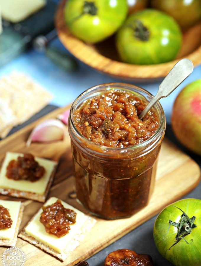 Jar of Easy Green Tomato Chutney with teaspoon inserted, on a board with cheese & crackers, surrounded by ingredients.