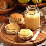 Easy Quince Curd in a jar (with teaspoon) on a board with buttered scones topped with curd, knife and quinces in a wooden bowl in background and more scones..