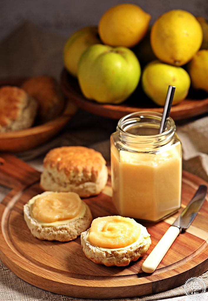 Easy Quince Curd in a jar (with teaspoon) on a board with scones, knife and quinces in a bowl.