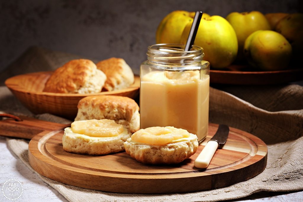 Easy Quince Curd in a jar (with teaspoon) on a board with scones, knife and quinces in a bowl.