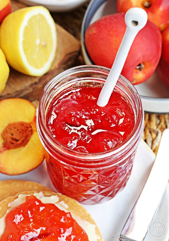 Open jar of Easy Nectarine Jam on a plate with Scotch Pancakes, cut lemon and a bowl of nectarines etc.