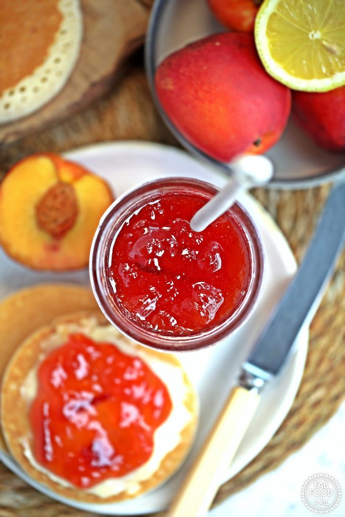 Overhead shot of open jar of Easy Nectarine Jam on a plate with Scotch Pancakes and a bowl of nectarines etc.