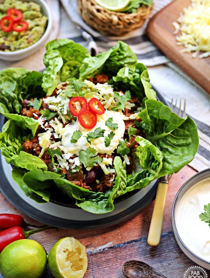 Side on portrait shot of Chilli Con Carne served in Little Gem lettuce leaves with grated cheese, a dish of sour cream, a dish of guacamole grated Cheddar cheese, limes, red chillies and a red pepper surrounding.