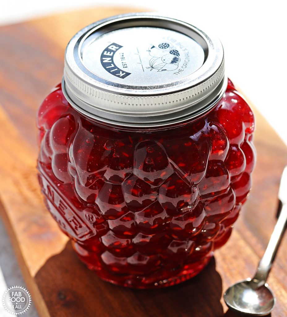 Jar of Quick & Easy Raspberry Jam (Kilner jar shaped like a raspberry) with it's lid on next to a silver spoon on a wooden board.