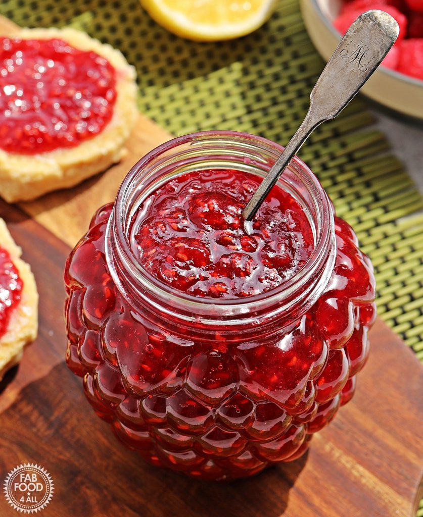 Quick & Easy Raspberry Jam with a silver spoon in it on a wooden board (scones, lemon half and bowl of raspberries just visible in the background.