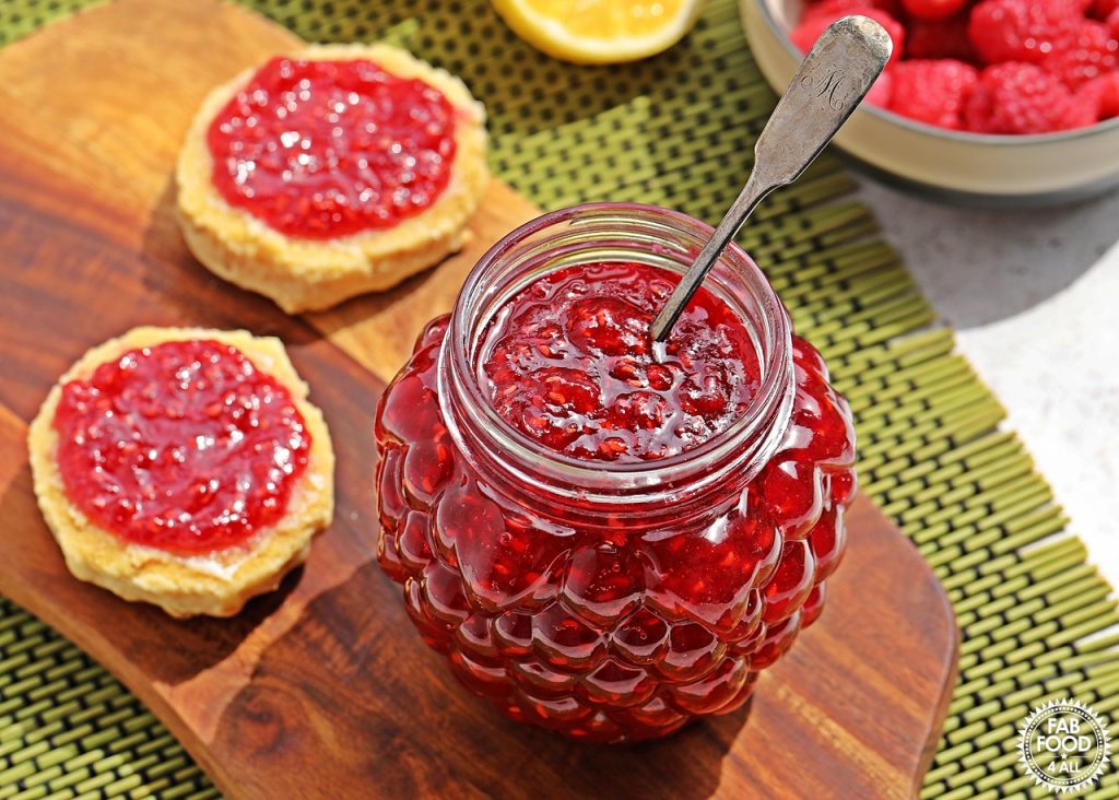 Open jar of homemade raspberry jam with a teaspoon in it (on a wooden board with 2 scone halves and the edge of a lemon and bowl of raspberries just visible in the background.