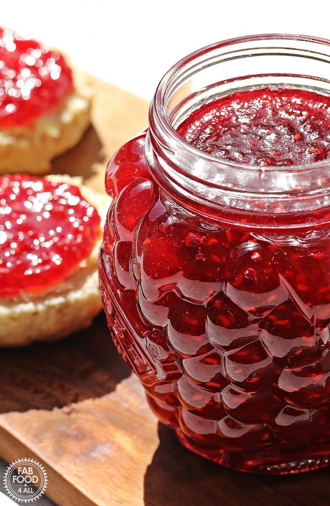 Jar of Quick & Easy Raspberry Jam on a wooden board with 2 scone halves with jam on in the background.