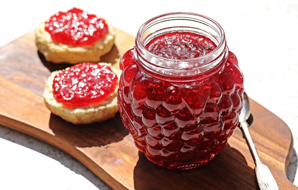 Quick & Easy Raspberry Jam on a board with 2 scone halves in the background with jam on.