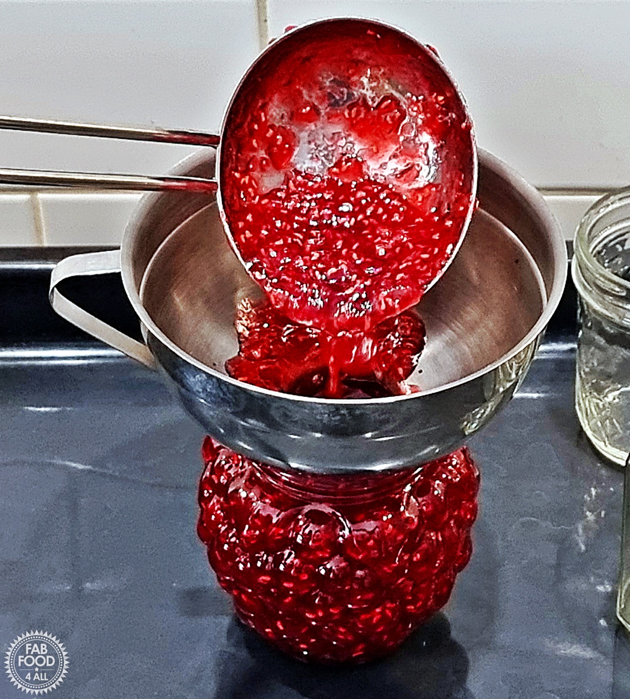 Hot raspberry jam being ladled into a jar with a jam funnel.