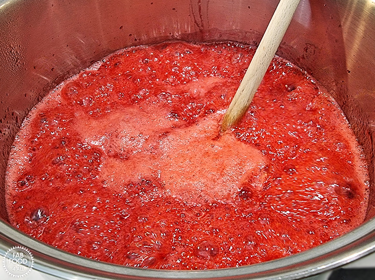 Raspberry jam on a rolling boil being stirred with a wooden spoon in a stainless steel pot.