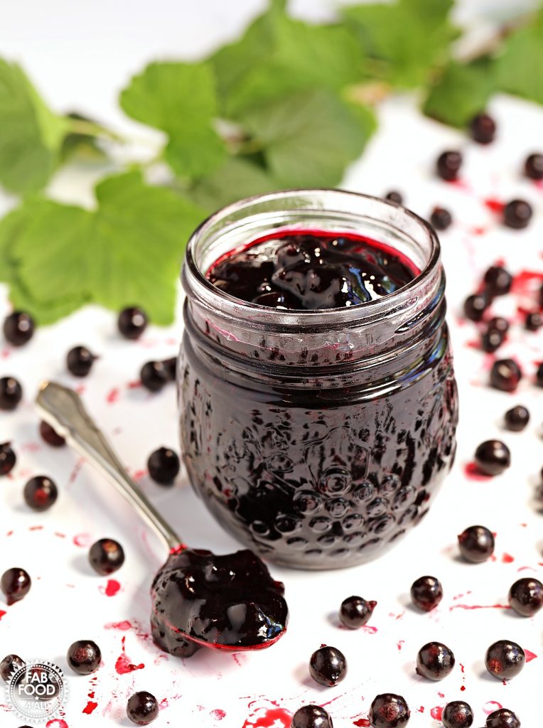 Pot of Easy Blackcurrant Jam on a white background with a teaspoon of jam next to it, blackcurrants scattered around it and blackcurrant leafy branch in the background.