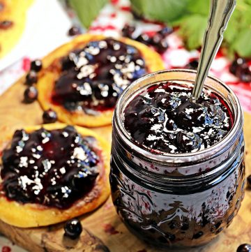 Easy Blackcurrant Jam in a jar with spoon on a board with 2 jam covered Lemon & Raisin Scotch Pancakes with blackcurrants dotted around and a leafy blackcurrant bush branch in the background.