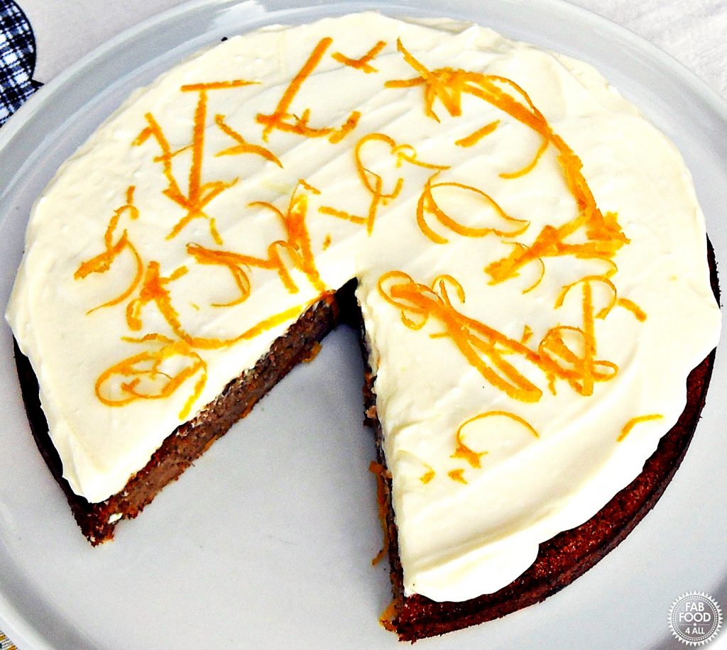 Quick & Easy Spelt Butternut Squash Cake with orange cream cheese frosting decorated in orange skin shavings on a white serving dish with slice cut out.