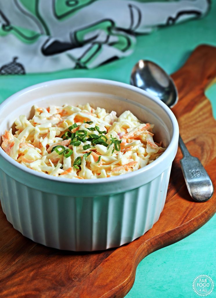 Bowl of coleslaw on a wooden board with serving spoon and tea towel in distance.