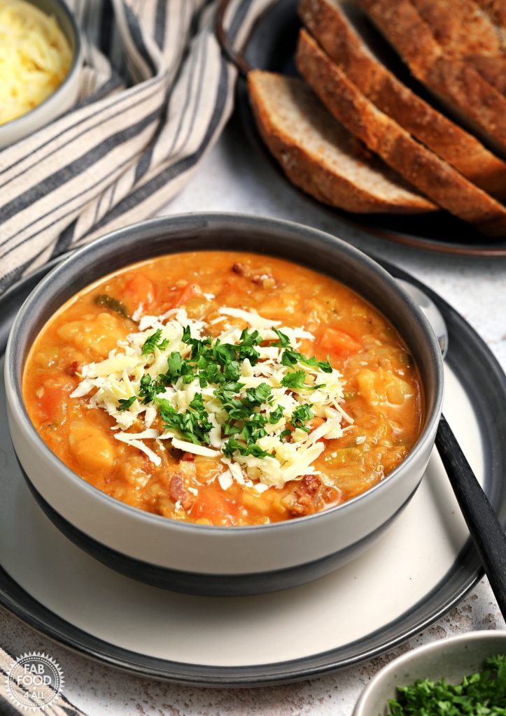 Glenda's Winter Lentil Soup in a bowl garnished with grated Cheddar cheese and chopped parsley. A tin platter of sliced crusty bread in the background and a bowl of grated Cheddar.