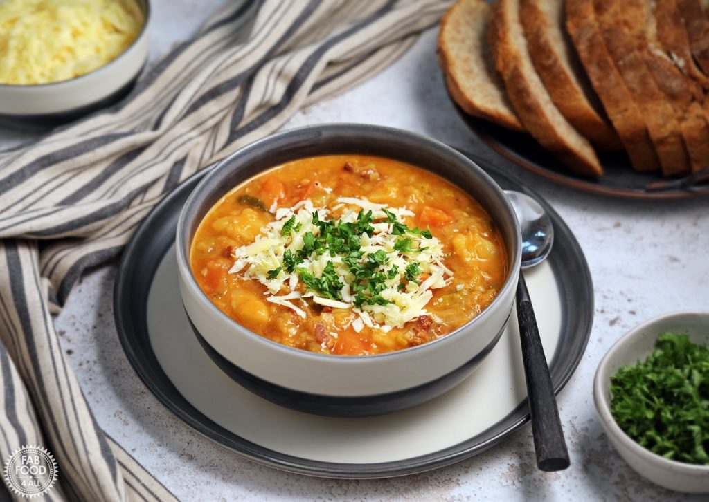 Landscape shot of Glenda's Winter Lentil Soup in a bowl garnished with grated Cheddar cheese and chopped parsley. A tin platter of sliced crusty bread in the background and a bowl of grated Cheddar.