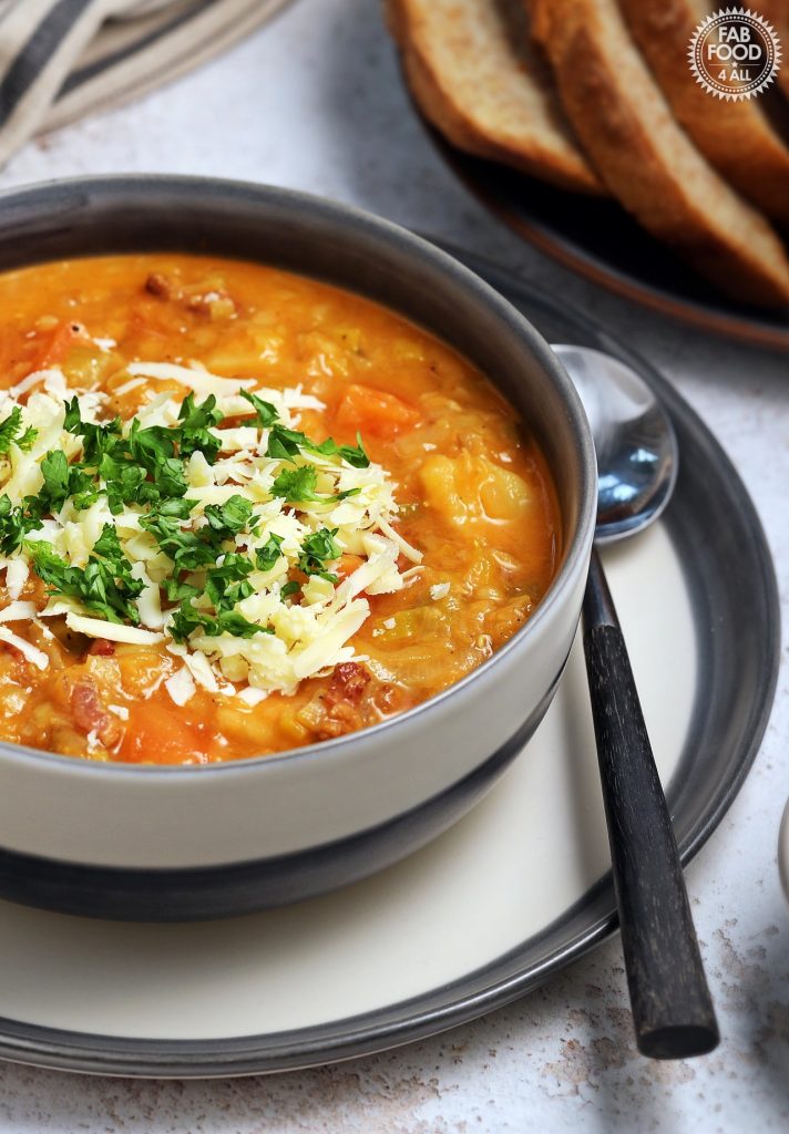 Close up of Landscape shot of Glenda's Winter Lentil Soup in a bowl garnished with grated Cheddar cheese and chopped parsley. A tin platter of sliced crusty bread in the background and a bowl of grated Cheddar.