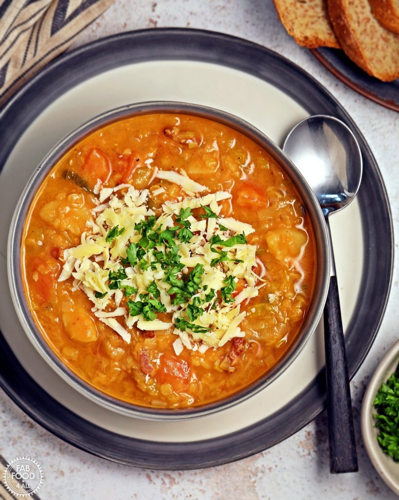 Close up overhead shot of Glenda's Winter Lentil Soup in a bowl garnished with grated Cheddar cheese and chopped parsley. A chunky soup made with carrots, parsnip, leeks, streaky bacon and orange juice.