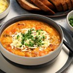 Portrait shot of Glenda's Winter Lentil Soup in a bowl garnished with grated Cheddar cheese and chopped parsley. A bowl of chopped parsley, grated cheese and platter of crusty bread in the background.