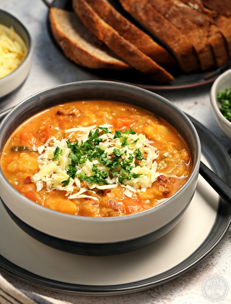 Portrait shot of Glenda's Winter Lentil Soup in a bowl garnished with grated Cheddar cheese and chopped parsley. A bowl of chopped parsley, grated cheese and platter of crusty bread in the background.