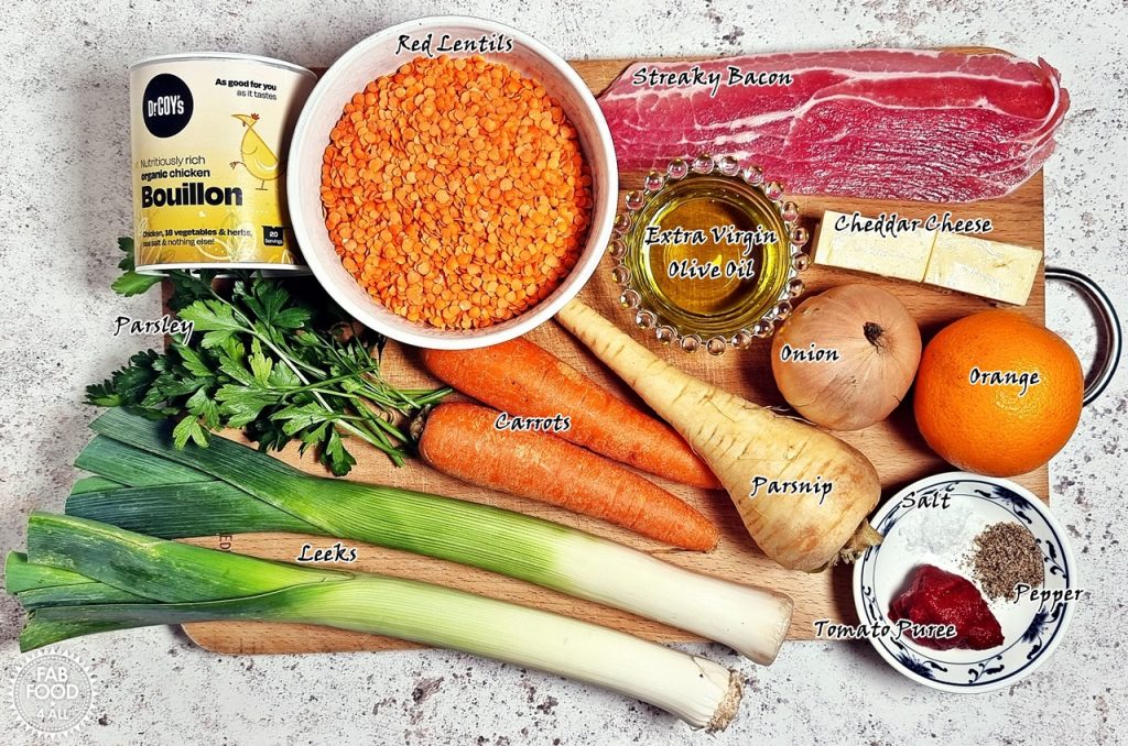 A wooden board with all the ingredients for Glenda's Winter Lentil Soup on it. Including Red Lentils, streaky bacon, bouillon, leeks, carrots, parsnip, orange etc.