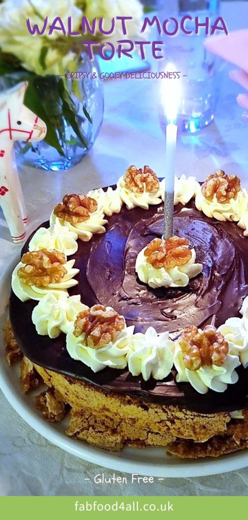 Walnut Mocha Torte on a platter with lit birthday candle. Plates, napkins and a vase of flowers in the background. Pinterest image.