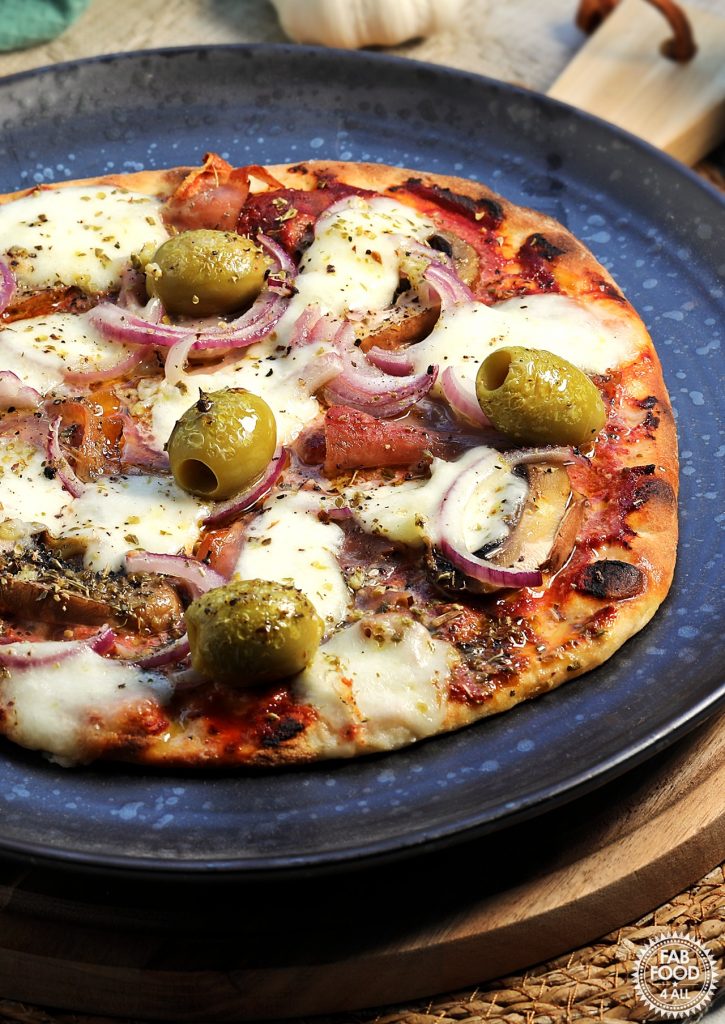 Easy Spelt Yogurt Flatbread Pizza (mushroom, Parma ham, big green olives, red onion and mozzarella on a tomato puree base) on a black plate on a round wooden board.