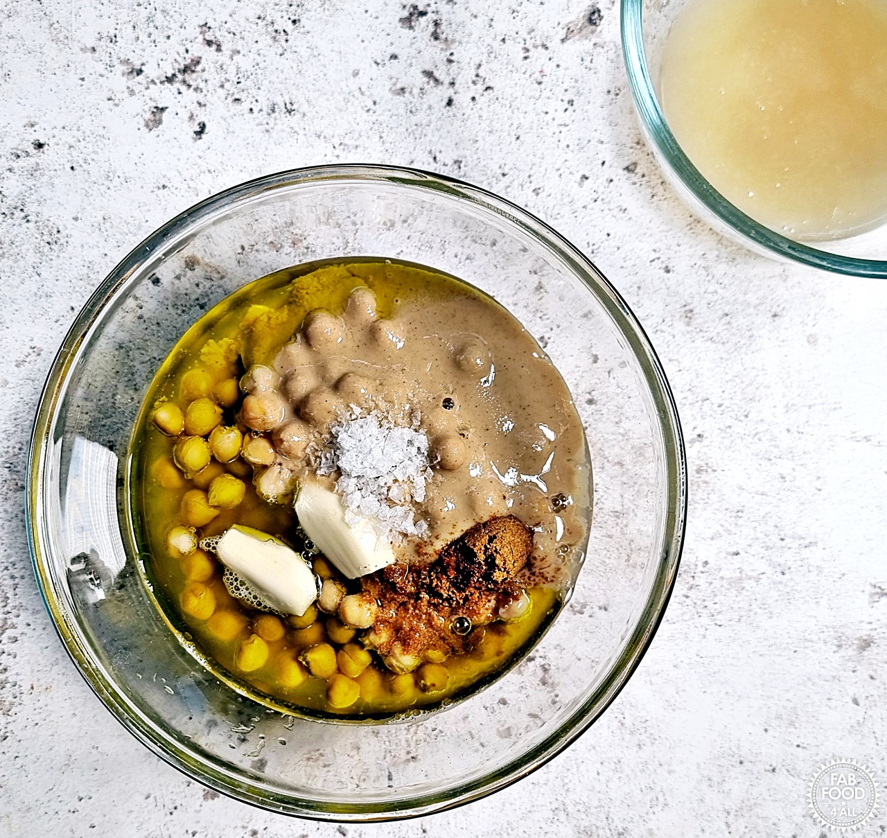 Hummus recipe ingredients in a glass bowl (chickpeas, lemon juice, EVOO, tahini, garlic, cumin, salt) with bowl of aquafaba just visible.