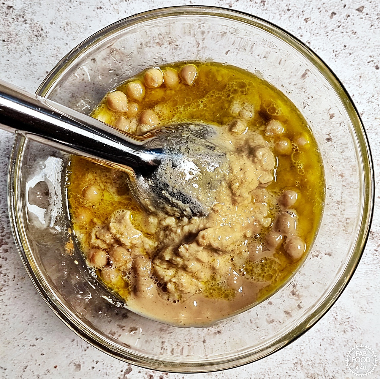Hummus ingredients being blended with a stick blender in a glass bowl.