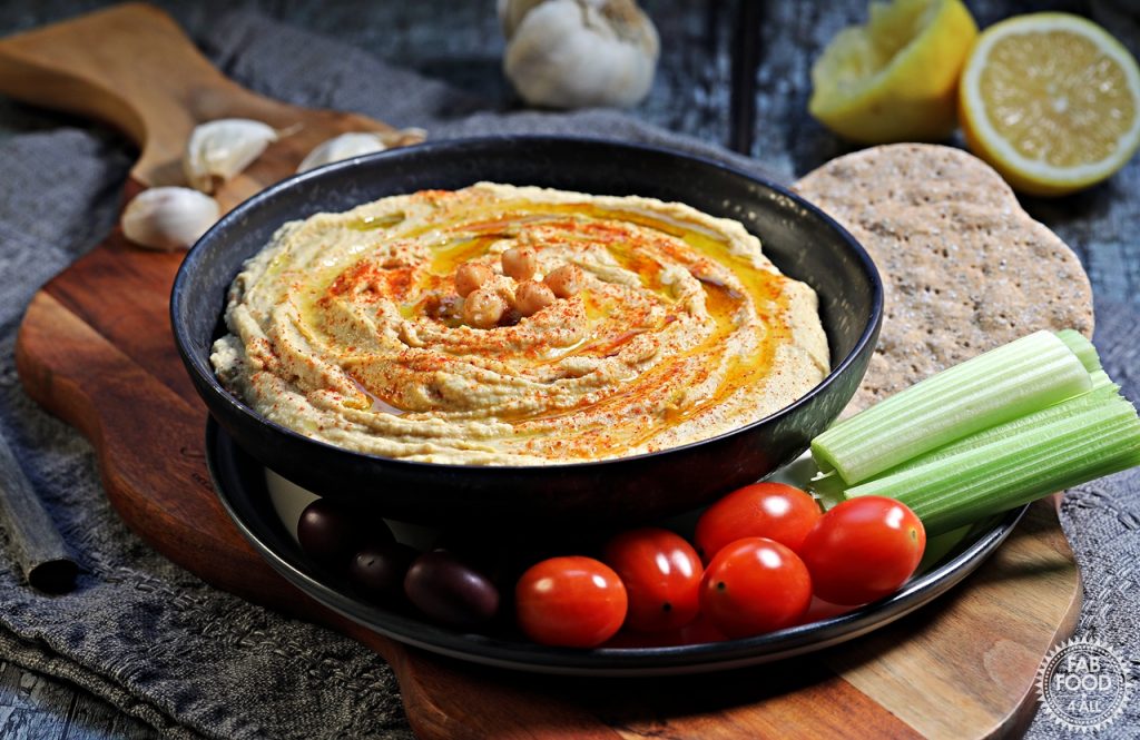 Landscape shot of hummus in a black dish surrounded by crudites and crackers. Garlic & lemon in the background.