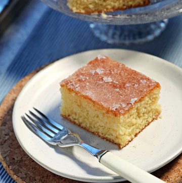 Close up of a slice of Lemon Drizzle Traybake on a plate with a glass pedestal with more slices in the background.