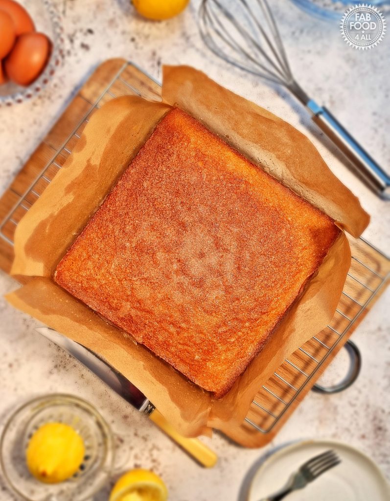 Overhead shot of Lemon Drizzle Traybake in parchment paper on a wire rack on a wooden board surrounded by a whisk, lemon juicer, eggs, a knife etc.