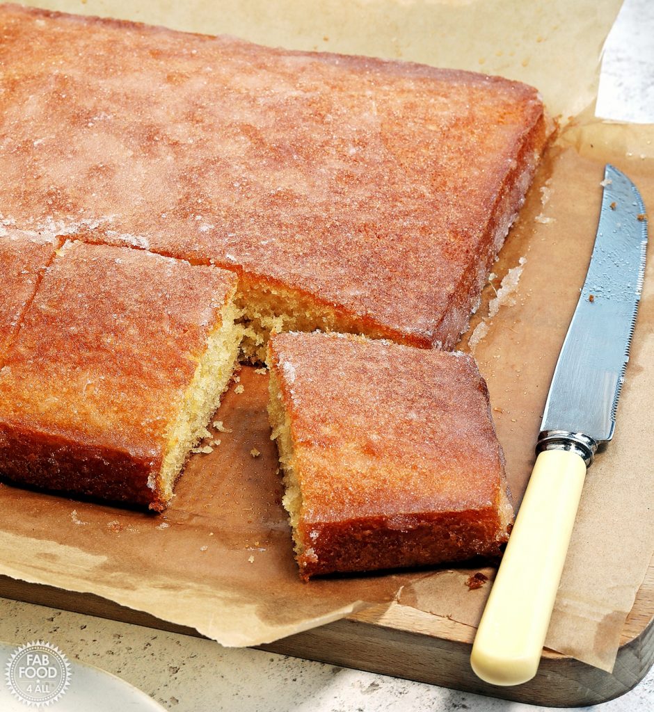 Lemon Drizzle Traybake cake with 2 slices cut from a corner. Shown on a board still in baking parchment with a cake knife.
