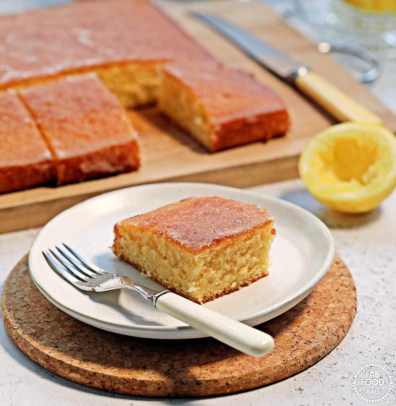 Close up of a slice of Lemon Drizzle Traybake on a plate with a fork on a cork mat. Half a lemon in the background and board with cake in the background plus a cake knife.