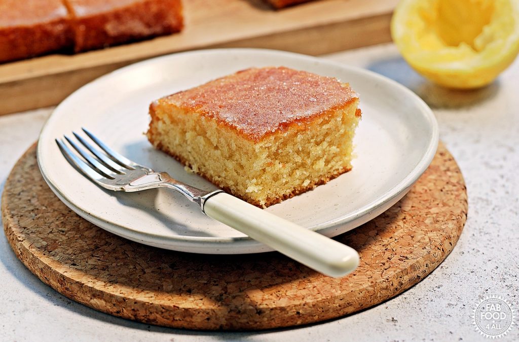 Close up of a slice of Lemon Drizzle Traybake on a plate with a fork on a cork mat. Half a lemon in the background and board with cake just visible.