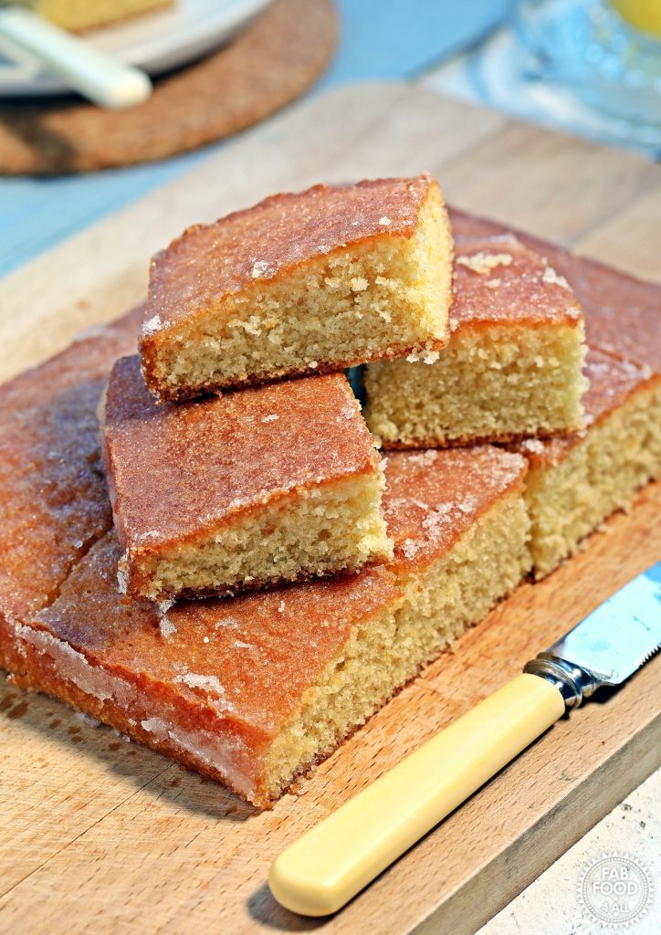 Lemon Drizzle Traybake slices stacked on top of teach other on a wooden board with knife showing in the foreground.