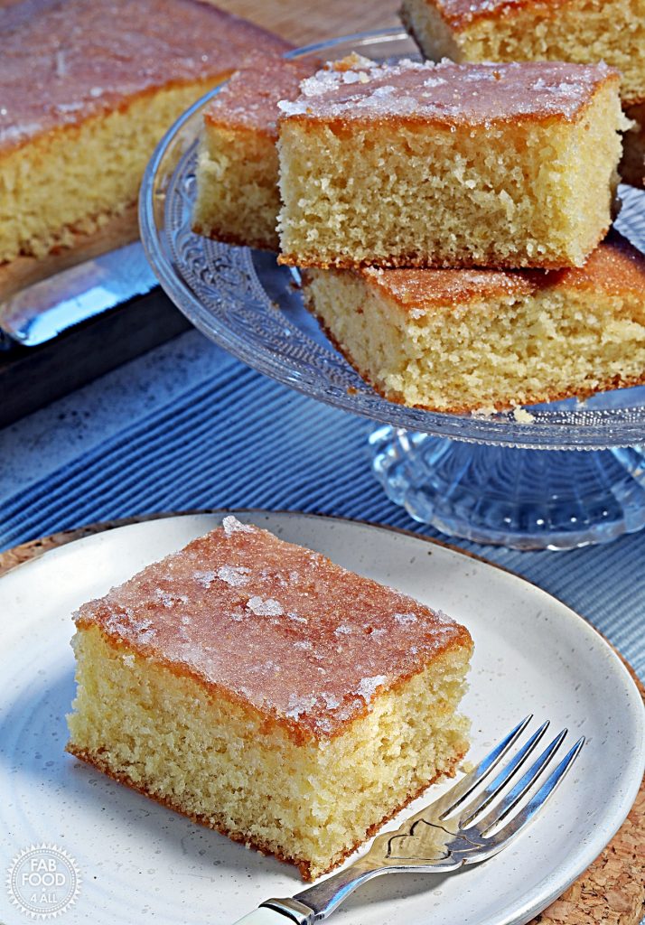 Close up of a slice of Lemon Drizzle Traybake on a plate with a glass pedestal with more slices and a board with the rest of the cake in the background.