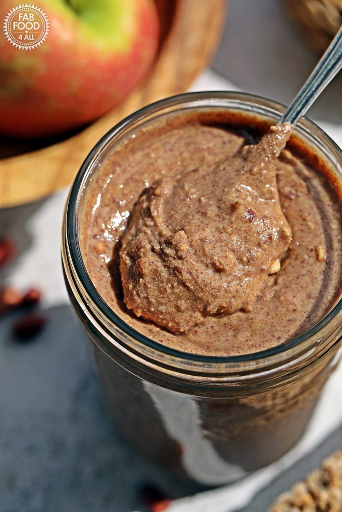 Jar of homemade crunchy peanut butter on a wooden board with a teaspoon of peanut butter sitting proud on top with bowl of apples just visible in background.