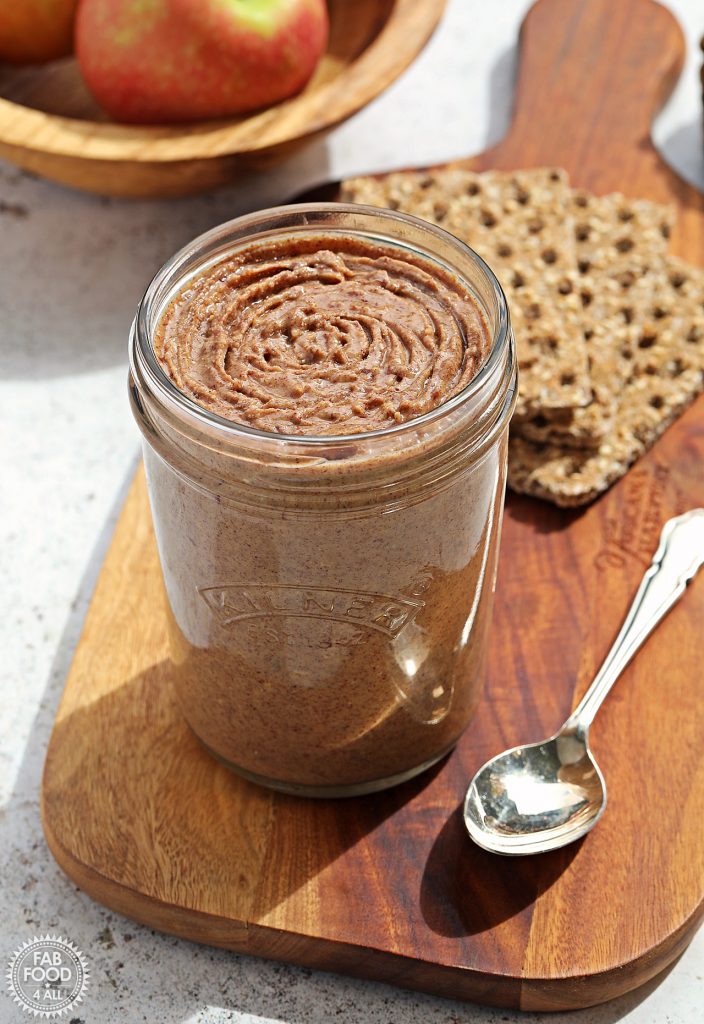 Open jar of Homemade Peanut Butter on a board with silver teaspoon, crackers and bowl of apples in background just visible.