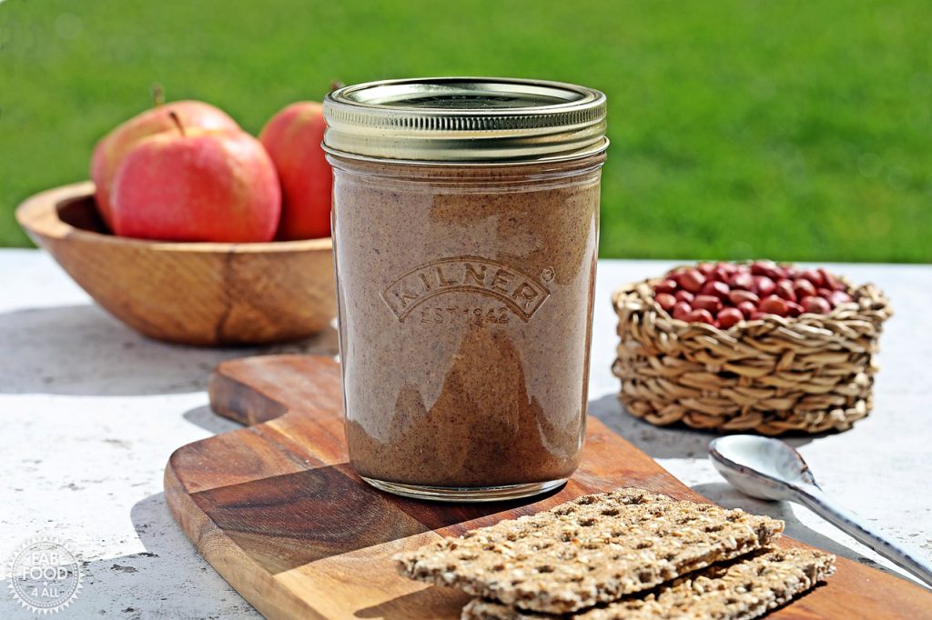 Jar of homemade peanut butter on a board with crackers and a wooden bowl of apples and a jute bowl of peanuts in the background.