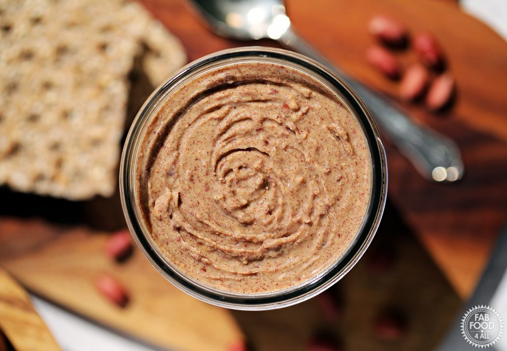 Overhead shot of jar of Homemade Peanut Butter on a board with crackers, teaspoon and a few red skin peanuts in the background.
