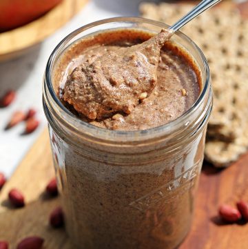 Jar of homemade crunchy peanut butter on a wooden board with a teaspoon of peanut butter sitting proud on top (surrounded by red skin peanuts with bowl of apples and crackers in the background).