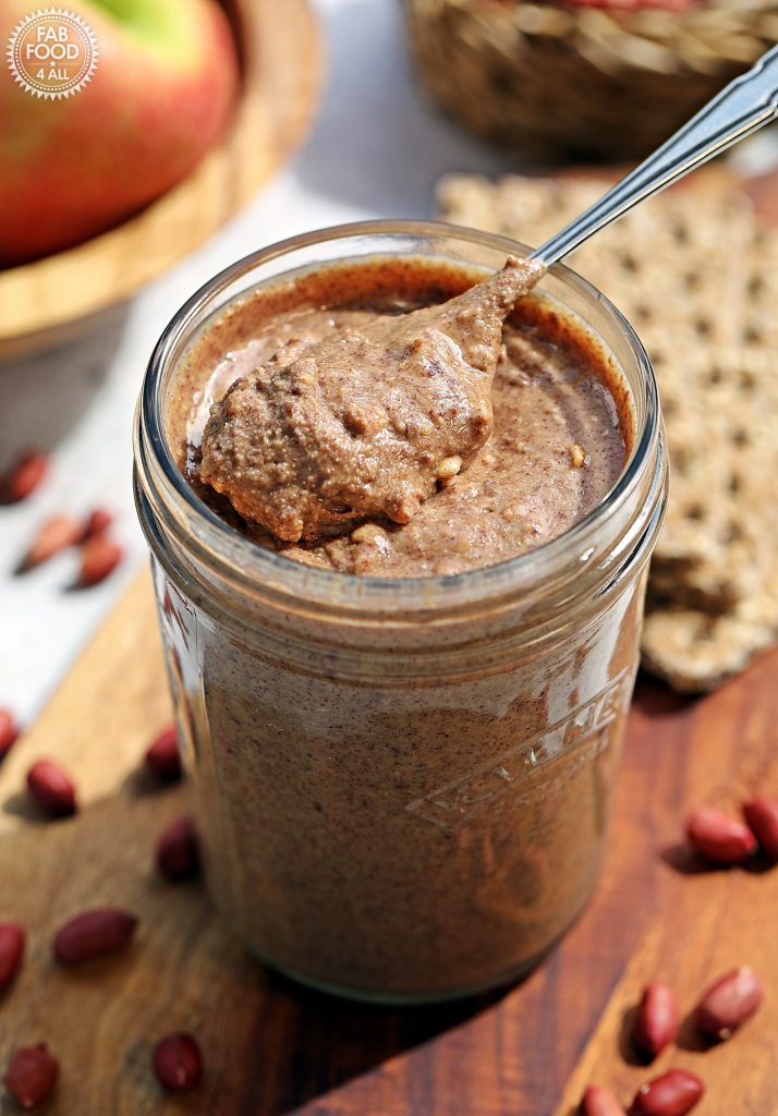 Jar of homemade crunchy peanut butter on a wooden board with a teaspoon of peanut butter sitting proud on top (surrounded by red skin peanuts with bowl of apples and crackers in the background).