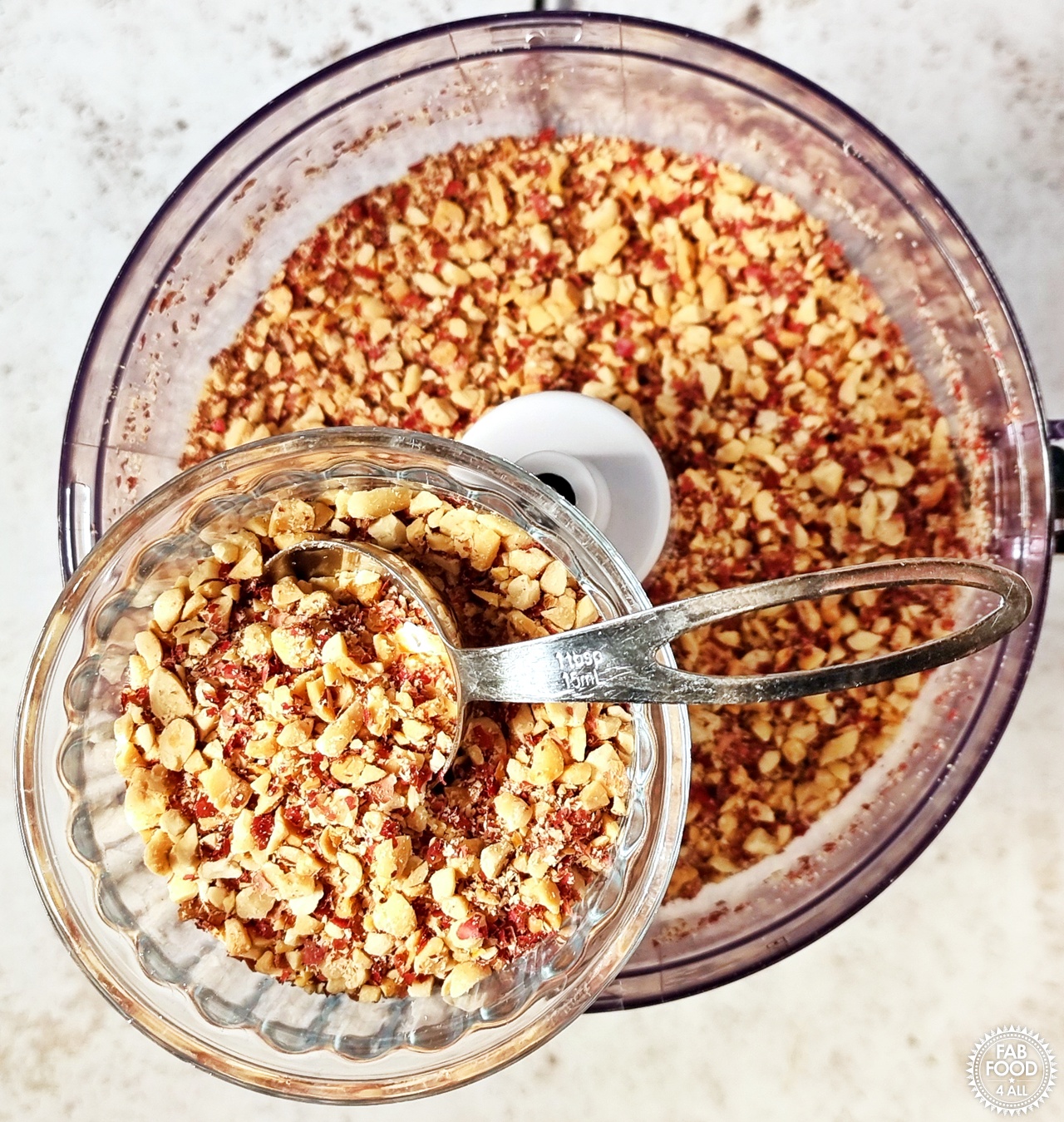 Ramekin of chopped nuts with tablespoon measure that have been removed from food processor in background below. (Optional, this is for crunchy peanut butter).