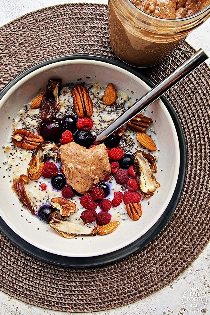 Bowl of porridge with mixed berries, nuts, chia seeds, dates and a dollop of homemade peanut butter with jar just visible behind.
