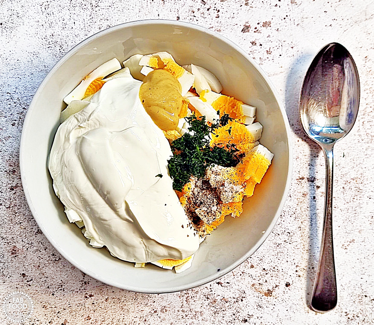 Egg Salad with Greek Yogurt ingredients (chopped boiled eggs, Greek yogurt, Dijon Mustard, chopped dill, sea salt flakes and freshly ground pepper) in a bowl with a spoon next to it.