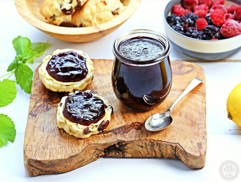 Pot of Blackberry & Raspberry Jam on top on a board with 2 scone halves (buttered and jammed) surrounded by raspberry stalks, a bowl of scones, a bowl or berries and a lemon.