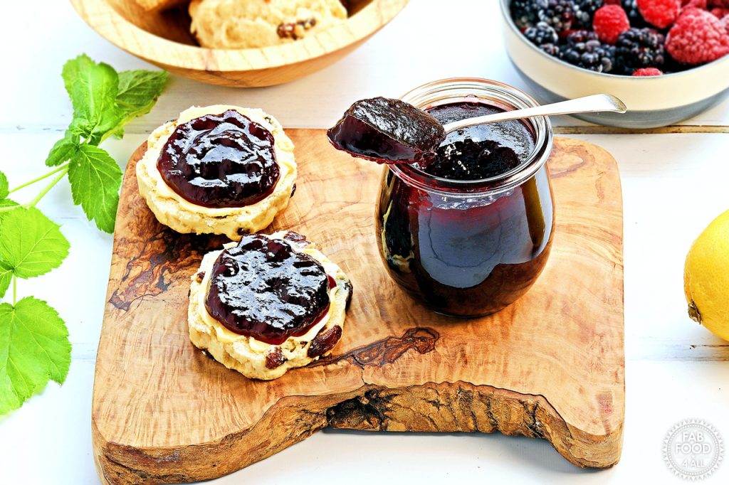 Blackberry & Raspberry Jam pot with a teaspoonful resting on top on a board with 2 scone halves (buttered and jammed) surrounded by raspberry stalks, a bowl of scones, a bowl or berries and a lemon.