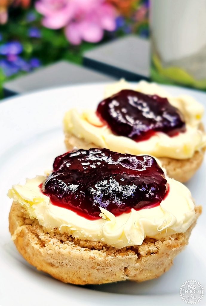 Plate of 2 Spelt Scone halves with clotted cream and Seedless Blackberry & Raspberry Jam on with mug and pink geranium just visible in the background.