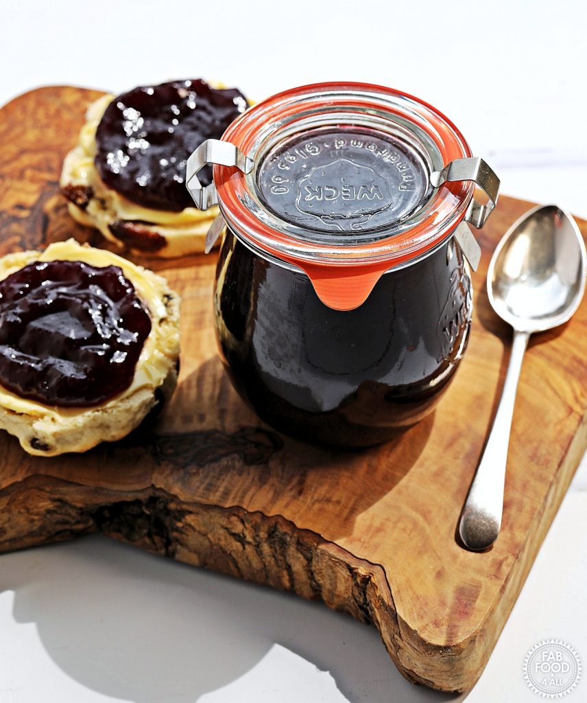 A Sealed Weck jar of Seedless Blackberry & Raspberry Jam on a board with a teaspoon and 2 scone halves with butter and jam on.